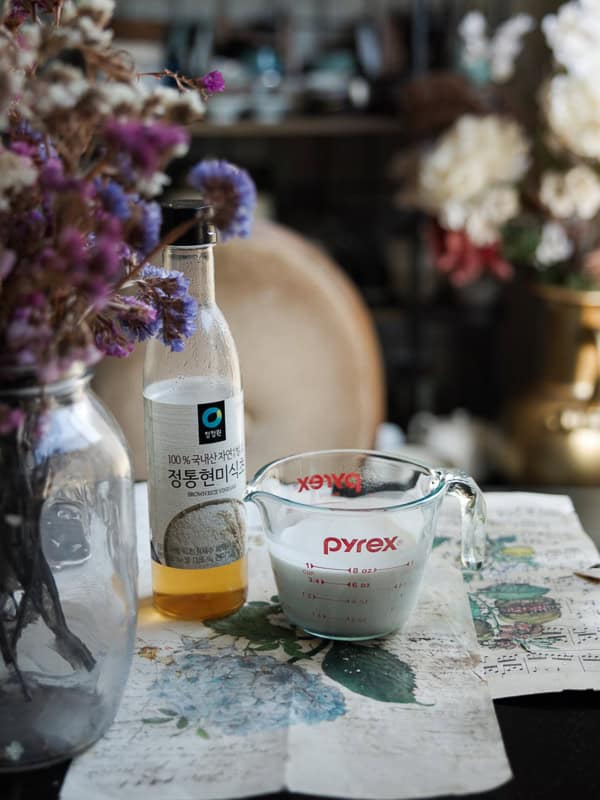 A glass bottle of rice vinegar stands beside a Pyrex measuring cup containing oat milk. They are on a table covered with floral-patterned paper, next to a vase of dried flowers. Blurred floral decor is visible in the background.